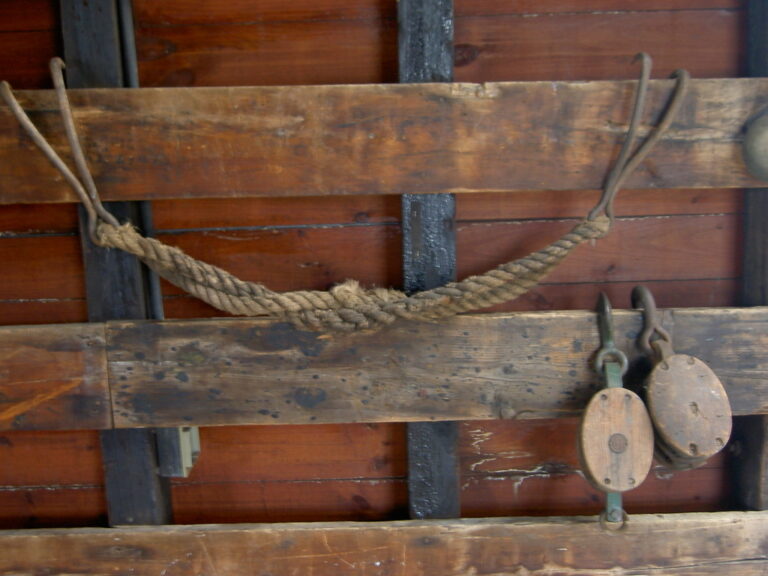 twisted arrangement of natural fiber rope slung between long shanked hooks suspended from a board on a wall near some blocks (pulleys)