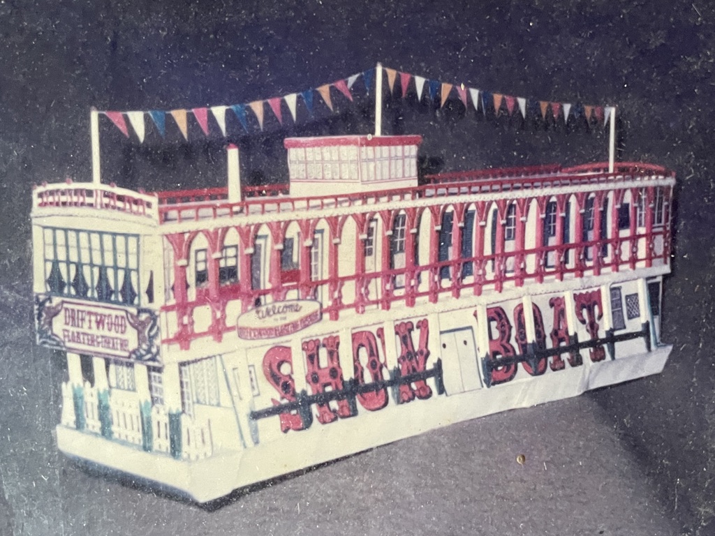 paper model of the Driftwood Theatre showboat barge, white with red trim, multicolor pennants strung over the top, and the words SHOW BOAT on the side in red